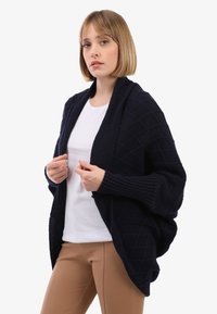 Marineblaue Strickjacke mit Rautenmuster, offener Vorderseite, gerippten Ärmeln und oversized Passform, kombiniert mit einem weißen Oberteil und beigen Hosen.
