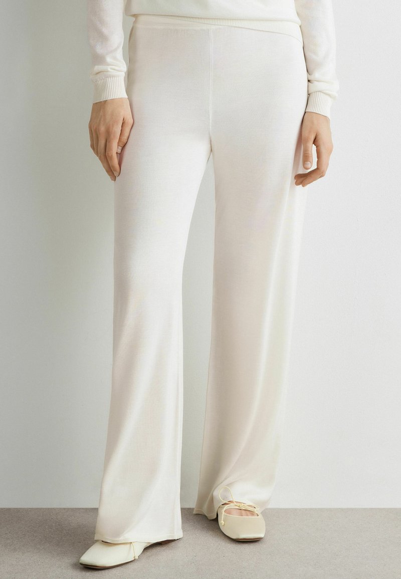 Persoon die losse, crèmekleurige broek draagt, gecombineerd met crèmekleurige platte schoenen met vierkante neus, staand op een lichtgrijze vloer tegen een witte muur.
