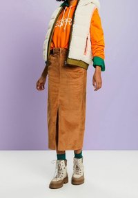 Brauner Cord-Midirock mit Schlitz vorne, kombiniert mit einem leuchtend orangefarbenen Hoodie und einer olivgrünen Steppweste. Cremefarbene Wanderschuhe.