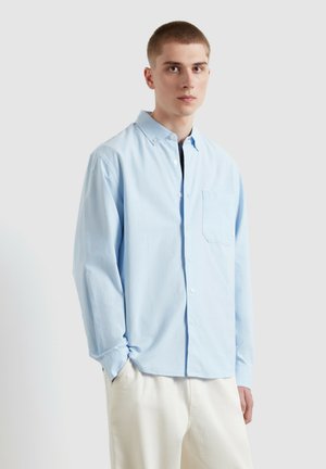 Giovane uomo con indosso una camicia celeste e pantaloni color crema, in piedi con una mano in tasca davanti a uno sfondo neutro.