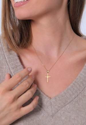 Frau trägt beige Strickpullover und berührt eine goldene Kreuzanhänger-Halskette auf ihrem Schlüsselbein, lächelt mit leicht geöffneten Lippen.