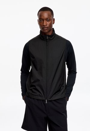 Jeune homme portant un gilet sans manches noir zippé par-dessus un tee-shirt noir à manches longues et un short noir, debout les mains dans les poches.