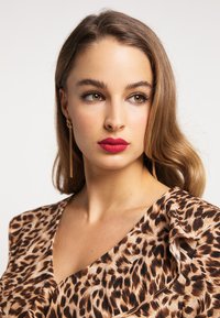 Bluse med leopardprint og rynket halsudskæring, lavet af letvægtsstof. Modellen har langt hår og bærer guld-dråbeøreringe. Lysende røde læber.