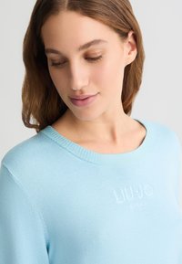 Maglione azzurro chiaro con scollo a giro ribattuto e logo ricamato discreto sul petto, realizzato in morbido materiale testurizzato.