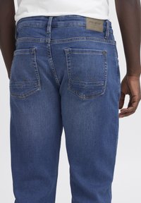 Kék vékony farmernadrág egyenes szabással, két hátul zsebbel, amelyek cikk-cakk varrással díszítettek, valamint márkás bőrpánttal a deréknál.