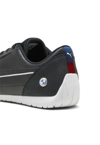 Baskets gris foncé avec une tige texturée, semelle blanche et accent bleu-rouge au niveau du talon. Présente un logo circulaire sur le côté.