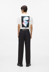 Cilvēks, kas valkā baltu t-kreklu ar izkropļotas sejas dizainu un tekstu "KARL LAGERFELD JEANS", melnas bikses, melnu jostu un melnus apavus.