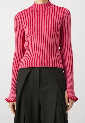 Femme portant un pull ajusté à manches longues, rayé verticalement de rose et rouge avec des poignets à volants, associé à un pantalon noir taille haute.