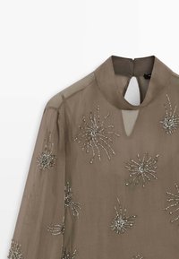 Gennemsigtig taupe kjole med høj krave, prydet med blomstermønstre af perler på forsiden og en nøglehulsåbning bagpå.