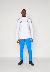 Bela športna majica z dolgimi rokavi z modrimi poudarki in zadrgo, kombinirana z modrimi hlačami. Na prsih je logotip Real Madrida, ob strani pa so črte.