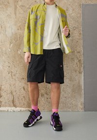 Camicetta a maniche corte con motivo a spirale verde e grigio, t-shirt crema, pantaloni cargo neri e sneaker nere/purple con calzini rosa.