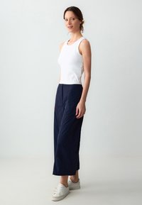 Witte mouwloze top gecombineerd met marineblauwe wijde broek. De outfit heeft een gladde textuur en een eenvoudig ontwerp, aangevuld met witte sneakers.