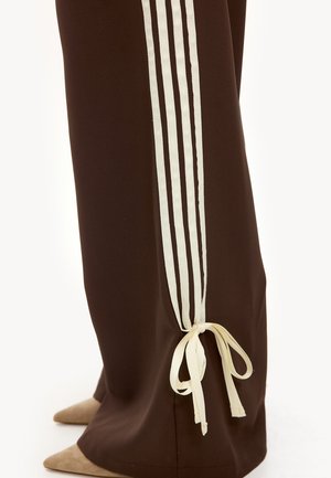 Bruine wijde broek met drie verticale witte strepen en een gestrikte witte band bij de enkel, gecombineerd met beige puntige schoenen.