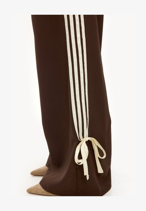 Pantalons évasés marron avec trois rayures blanches verticales et un ruban blanc noué près de la cheville, associés à des chaussures beige à bouts pointus.