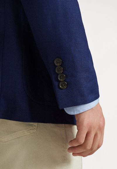 Mužská ruka vedle rukávu tmavě modrého saka se čtyřmi černými knoflíky, manžetou světle modré vzorované košile a viditelným kapesníkem béžových kalhot.