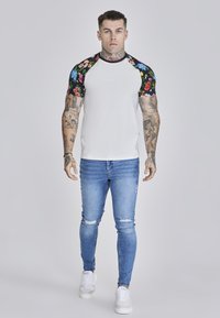White t-shirt with floral black sleeves, blue distressed skinny jeans, and white sneakers worn by a tattooed man. Simple, bold design.