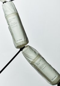 To sylindriske kroppsserumflasker pakket inn i hvitt papir, festet med en svart snor, med minimalistiske etiketter med tekst.