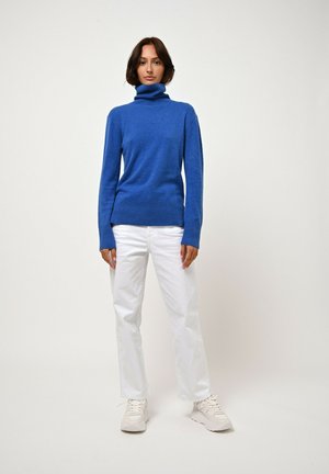 Maglione a collo alto blu reale, con texture morbida, abbinato a jeans bianchi a gamba dritta e sneakers chiare, in piedi davanti a uno sfondo neutro.