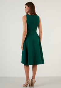 Ärmlös grön klänning med en figurnära bodice och utsvängd kjol, med en dragkedja i ryggen och slät tygtextur. Kjolens fållar slutar strax ovanför knäet.