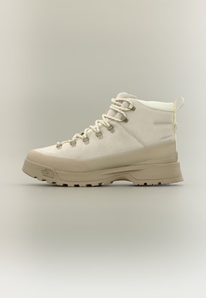 Botte de randonnée beige avec semelle épaisse, lacets à l'avant, col rembourré et languette à l'arrière, présentée sur un fond neutre.