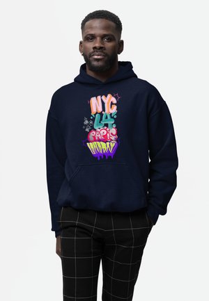 Mann trägt schwarz-weiß kariere Hosen und einen marineblauen Hoodie mit buntem Graffiti-Text, auf dem "NYC", "LA", "Paris" und "London" steht.