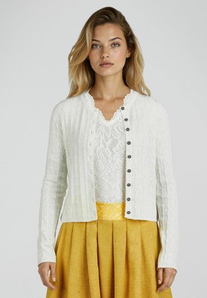 Woman wearing a white lace top with a white buttoned cardigan and a pleated mustard yellow skirt with a floral waistband.