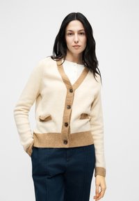 Γυναίκα με μακριά σκουρόχρωμα μαλλιά που φοράει μπεζ ζακέτα με καφέ λεπτομέρειες και σκούρο μπλε παντελόνια, στέκεται μπροστά σε απλό ανοιχτόχρωμο φόντο.