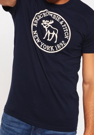 Marineblå T-shirt med hvidt broderet Abercrombie & Fitch-logo, der viser en elg og teksten "New York 1892" på brystet.