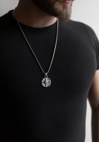 Pendentif en argent en forme de boussole sur une chaîne métallique, orné de points cardinaux et d'une gemme centrale, porté sur un T-shirt noir ajusté.