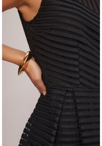 Czarna, strukturalna sukienka z pionowymi paskami. Lśniąca złota bransoletka noszona na nadgarstku, podkreślająca elegancki sylwetkę.