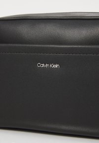 Black synthetic leather bag with a smooth texture. Features a silver "Calvin Klein" logo and subtle stitching detail. Rectangular shape.