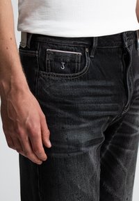 Nahaufnahme einer Hand einer Person neben schwarzen Jeans mit einem kleinen gestickten "J" an der Münztasche, die ein weißes Hemd trägt.