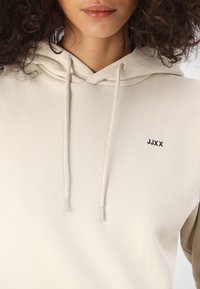 Bēša kapuce ar auklu, mīksta tekstūra, neliels melns "JJXX" logo uz krūšu un kontrastējošas piedurknes tumšākā tonī.