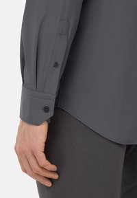 Camisa gris de manga larga con puños abotonados, hecha de una tela suave. El puño tiene dos botones y un detalle de diseño puntiagudo.
