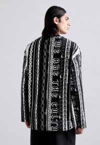 Čierno-biela košeľa s dlhým rukávom, ktorá obsahuje grafický vzor reťazí a textu, uvoľneného strihu a hladkej textúry látky.