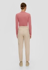 Femme avec les cheveux en chignon portant un haut rose à manches longues rentré dans un pantalon beige et des bottines beige, se tenant devant un fond uni.