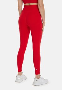 Leggings rouges en tissu extensible avec une taille haute et une texture lisse. Détail notable : petit logo blanc sur la jambe inférieure.