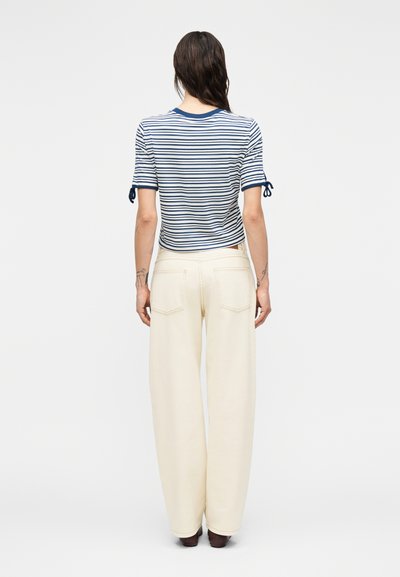 Ριγέ τοπ με κοντά μανίκια σε μπλε και λευκές οριζόντιες ρίγες, συνδυασμένο με κρεμ παντελόνια με φαρδιά μπατζάκια. Το τοπ διαθέτει κορδόνια στα μανίκια.