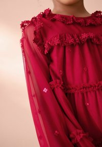Bambina che indossa un vestito rosso trasparente con ricami floreali e dettagli a ruches sul colletto, sulle maniche e in vita, su uno sfondo neutro.