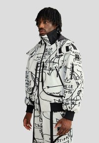 Hőszigetelt fehér dzseki fekete absztrakt grafikákkal, cipzáras zsebekkel, bordázott mandzsettákkal és magas gallérral, hozzáillő nadrággal párosítva.