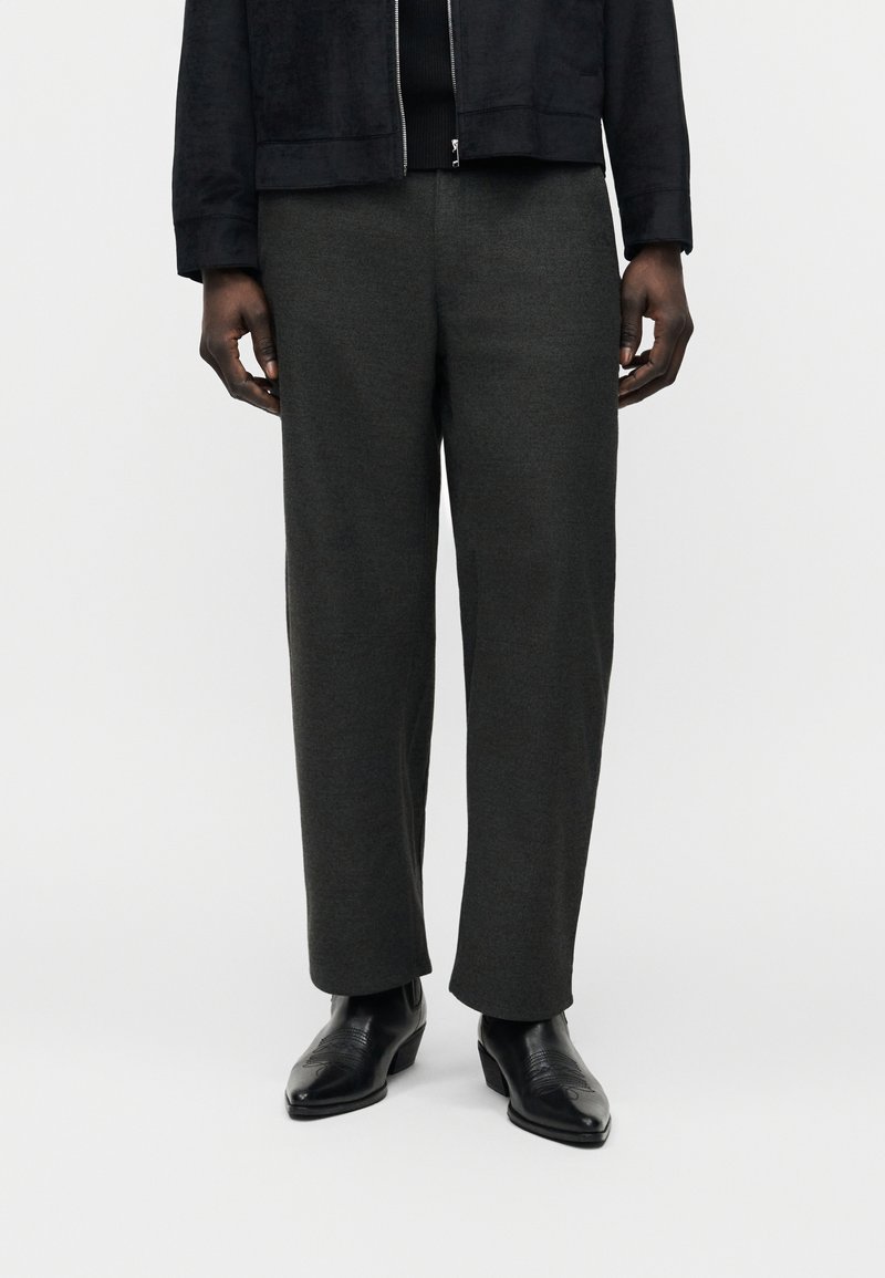 Homme portant un pantalon droit gris foncé, des bottes en cuir noires et une veste zippée noire, debout devant un fond blanc.