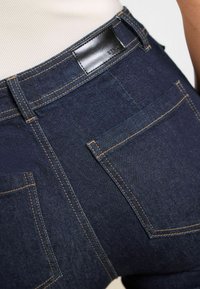 Mörkblå denimjeans med bruna sömmar, med en läderetikett i midjan och två bakfickor. Slät textur och åtsittande design.