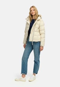 Pufferjack in crème met een ritssluiting aan de voorkant, capuchon en een gewatteerd patroon. Gecombineerd met blauwe jeans en witte sneakers, op een effen achtergrond.