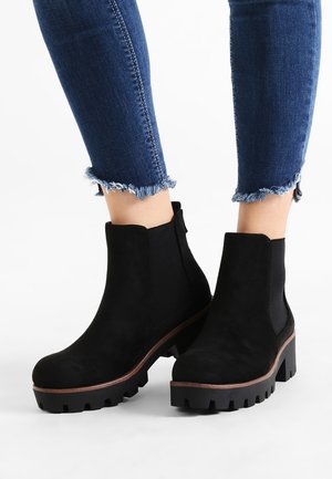 Bottines en daim noir avec une semelle épaisse, présentant un design à enfiler et un talon en bois épais. La texture est lisse avec des coutures prononcées.