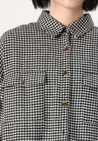 Black and white houndstooth shirt with a collared neck, two front pockets, and brown buttons on a textured fabric.