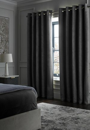 Dark gray blackout curtains partly open on a window in a modern bedroom with a bed, nightstand, lamp, and textured rug.