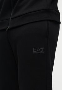 Melnas sporta bikses no mīksta auduma ar kabatu ar rāvējslēdzēju un EA7 logotipu uz augšstilba. Gluda tekstūra un piegulošs dizains.