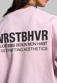 Růžová bavlněná mikina s velkým černým textem na zadní straně. Text obsahuje "NRSTBHVR" a "BERLIN MON HABIT", s uvolněným střihem a kulatým výstřihem.