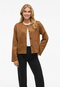 Veste courte en daim marron avec un col rond, fermeture par bouton central et poches avant. Portée sur un haut blanc et associée à un pantalon noir.