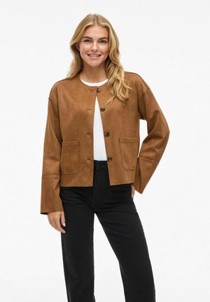 Veste courte en daim marron avec un col rond, fermeture par bouton central et poches avant. Portée sur un haut blanc et associée à un pantalon noir.